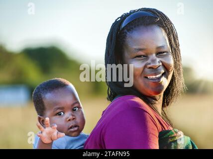 afrikanische Dorfmutter mit Zöpfen trägt das Kind in eine Decke gewickelt im Rücken Stockfoto