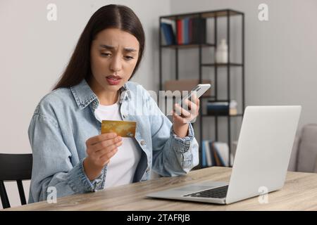 Verwirrte Frau mit Kreditkarte, Smartphone und Laptop am Tisch drinnen. Seien Sie vorsichtig – Betrug Stockfoto