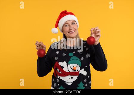 Glückliche ältere Frau im Weihnachtspullover und Weihnachtsmann-Hut mit festlichen Kugeln auf orangem Hintergrund Stockfoto