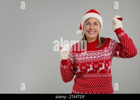 Glückliche Seniorin in Weihnachtspullover, Weihnachtsmütze und gestrickten Fäustlingen auf grauem Hintergrund. Leerzeichen für Text Stockfoto