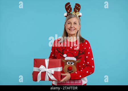 Ältere Frau im Weihnachtspullover und Rentier-Stirnband, das Geschenk auf hellblauem Hintergrund hält Stockfoto