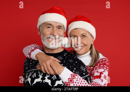 Älteres Paar in Weihnachtspullover und Weihnachtsmannmützen auf rotem Hintergrund Stockfoto