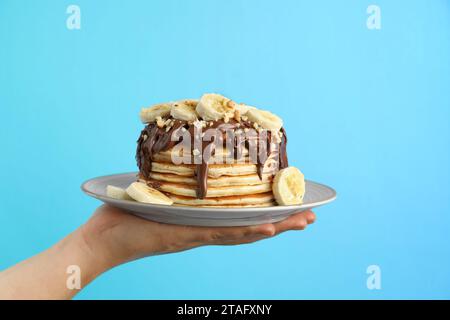 Frau, die einen Teller mit leckeren Pfannkuchen mit Schokoladenaufstrich hält, Bananenscheiben und Nüssen auf hellblauem Hintergrund, Nahaufnahme Stockfoto