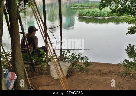 Ein einsamer Fischer sitzt im Schatten am Ufer eines ruhigen Sees in Hanoi, Vietnam. Diese städtischen Wasserstraßen beherbergen eine Vielzahl von Süßwasserfischen Stockfoto