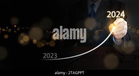 Ein Geschäftsmann zeichnet auf virtuellen technischen Bildschirmen eine steigende Kurve, von Ende 2023 bis Anfang 2024. Wir freuen uns über ein frohes neues Jahr und freuen uns auf ein neues Geschäftsziel Stockfoto