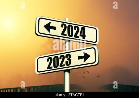 Straßenschilder zeigen den Weg zum neuen Jahr 2024 und zum alten Jahr 2023 an einem blauen Himmel mit Nebel und Sonnenuntergang, kreative Idee. Wählen Sie eine neue Straße, ein neues Konzept. Stockfoto