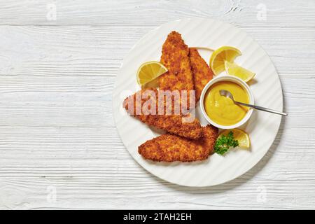 Im Ofen gebackenes paniertes Fischfilet mit gelbem Senf und Zitronenscheiben auf weißem Teller auf weißem Holztisch, flach gelegen, freier Platz Stockfoto