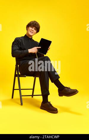 Junger attraktiver Mann, Geschäftsmann, der auf einem Stuhl sitzt, hält ein Tablet und zeigt auf den leeren Bildschirm vor lebendigem gelbem Studiohintergrund. Stockfoto