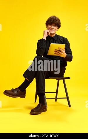 Junger attraktiver, fröhlicher Mann, Geschäftsmann, der auf Stuhl sitzt, hält Tablet und blickt auf die Kamera vor lebendigem gelbem Studiohintergrund. Stockfoto