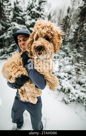 Ein lächelnder junger Mann hält seinen schneebedeckten Hund, während er in NH läuft Stockfoto