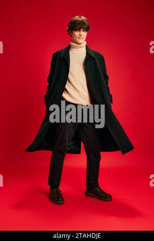 Porträt eines lächelnden jungen Mannes, der in einem modernen, lässigen, warmen Outfit vor rotem Studiohintergrund posiert. Stockfoto