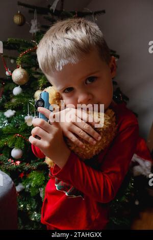 Ein Kind in Rot umarmt einen Teddybären und ein Auto in der Nähe eines weihnachtsbaumes Stockfoto