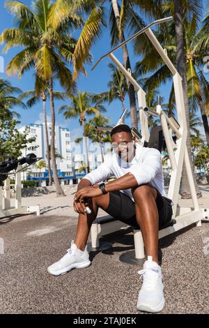 Glücklicher afroamerikanischer Sportler in schnurlosen Ohrhörern, der nach dem Training in Miami Beach ruht Stockfoto