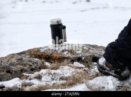 Halt im Winter. Der Reisende ruht sich auf dem moosigen Stein aus und trinkt heißen Tee aus einer Thermoskanne Stockfoto