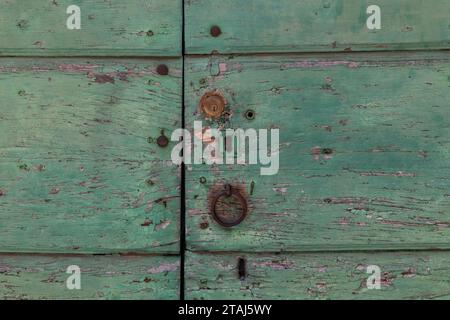 Eine verblasste, abblätternde und verwitterte, hellgrün lackierte Tür mit abgebrochener Oberfläche, Schlüssellöchern, Schloss und rostfreiem Metallklopfer. Stockfoto