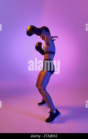 Wildes und selbstbewusstes, entschlossenes Boxermädchen, professioneller Sportler, der sich im violetten Neonlicht vor dem Hintergrund eines Farbstudios ausgibt. Konzept von Stockfoto