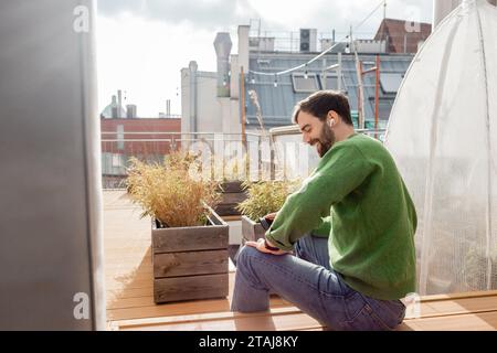Lächelnder bärtiger Mann in Ohrhörern und lässiger Kleidung mit Smartphone auf dem Dach der Hausterrasse Stockfoto