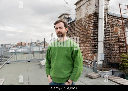 Bärtiger Mann, der auf der Dachterrasse des Hauses steht, mit verschwommenem Stadtbild im Hintergrund in Wien Stockfoto