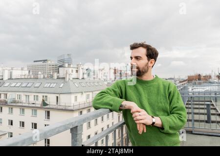 Tätowierter Mann im grünen Pullover, der wegblickt, während er auf der Dachterrasse in Wien steht Stockfoto