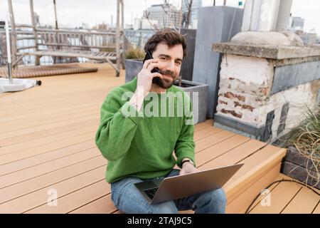 Bärtiger Mann, der auf dem Smartphone spricht und an einem Laptop auf der Dachterrasse in Wien arbeitet Stockfoto