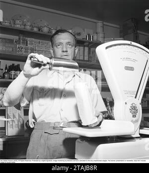 In einem Supermarkt in den 1940ern Ein Ladenbesitzer wird gesehen, wie er etwas in eine Papiertüte gießt. In den Regalen hinter ihm sind Pakete und Flaschen ordentlich aufgereiht. Eine weiße Ladenwaage steht auf dem Tresen und war ein unverzichtbarer Bestandteil des Geschäfts, da viele Waren in großen Mengen verkauft wurden. 1947. Kristoffersson Ref. AC127-4 Stockfoto