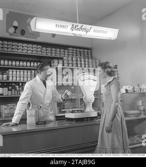 Frau in einem Lebensmittelgeschäft in den 1940ern Eine junge Frau steht an der Theke, wo ein Händler ein Glas Rhabarbercreme hält. In den Regalen hinter ihm sind Pakete und Flaschen ordentlich aufgereiht. Eine weiße Ladenwaage steht auf dem Tresen und war ein unverzichtbarer Bestandteil des Geschäfts, da viele Waren in großen Mengen verkauft wurden. Schweden 1949. Kristoffersson Ref. AP72-5 Stockfoto