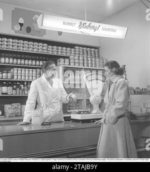 Frau in einem Lebensmittelgeschäft in den 1940ern Eine junge Frau steht an der Theke, wo ein Händler ein Glas Rhabarbercreme hält. In den Regalen hinter ihm sind Pakete und Flaschen ordentlich aufgereiht. Eine weiße Ladenwaage steht auf dem Tresen und war ein unverzichtbarer Bestandteil des Geschäfts, da viele Waren in großen Mengen verkauft wurden. Schweden 1949. Kristoffersson Ref. AP72-5 Stockfoto