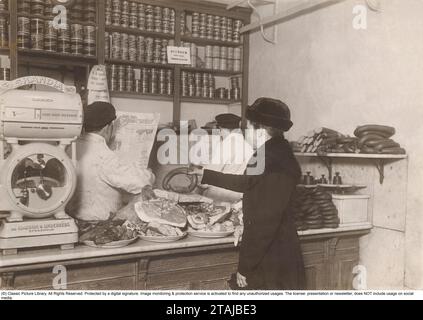 In den 1920er Jahren Eine Kundin in einem Lebensmittelgeschäft in Stockholm. Hier liegen sowohl Leber als auch Schweinefleisch und anderes Fleisch völlig offen auf der Theke. Auch ein Schweinekopf. Sie hat sich jedoch entschieden, eine geräucherte Wurst zu kaufen und sie dem scheinbar uninteressierten Disponenten zu überlassen. Er hat seine Augen woanders, nämlich in Torsten Tegnérs Zeitung Idrottsbladet. Schlagzeilen auf der Titelseite sind interessanter als die Wünsche des Kunden zu erfüllen. Der andere Disponent ist nicht viel besser und hat dem Kunden den Rücken zugewandt und liest eine Zeitung. Hier in den 1920er Jahren war der Umgang mit Fleisch völlig anders Stockfoto