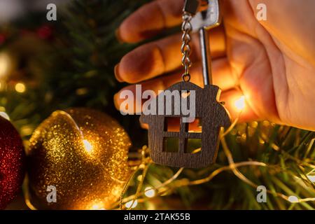 Geschenk Schlüsselhaus mit Schlüsselanhänger in der Hand Hintergrund des Weihnachtsbaums. Bauen, entwerfen, Projekt, Umzug in ein neues Haus, Hypothek, Miete und Kauf von Immobilien Stockfoto