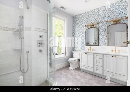 Ein gemütliches Badezimmer mit einer blauen Tapete, grauem Schrank, goldenem Wasserhahn und Spiegeln und einer begehbaren Dusche mit Fliesen. Stockfoto
