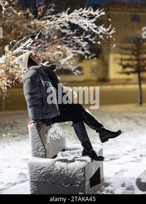 Junge Frau, die nachts im verschneiten Winterpark auf einer Bank sitzt, vor dem Hintergrund der Lichter der Stadt. Stockfoto