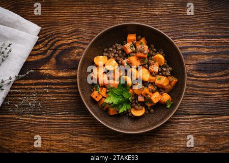 Gesunde vegane Mahlzeit, winterlicher warmer Salat mit gebratenen Süßkartoffeln, Linsen, goldenen Berris und Kräutern in Keramikschale auf rustikalem Holztisch, direkt Stockfoto