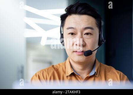 Nahaufnahme eines jungen asiatischen Mannes, der konzentriert und ernsthaft im Büro an einem Laptop mit Headset arbeitet. Stockfoto