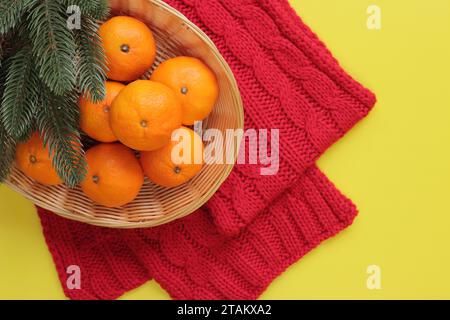 Mandarinen in einem Korb, ein roter Strickschal und Tannenzweige auf gelbem Hintergrund. Weihnachtskomposition, Neujahrseinrichtung Stockfoto