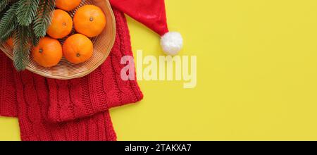 Mandarinen im Korb, roter Strickschal, Weihnachtsmann-Hut, Fichtenzweige auf gelbem Hintergrund. Weihnachtskomposition, Neujahr, Banner. Silvester-Deko Stockfoto