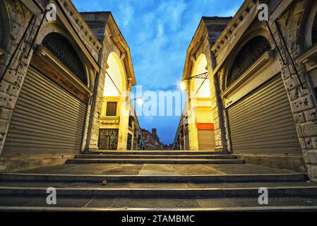 Die Rialto-Brücke in Venedig wird bei der ersten Ampel überquert, keine Menschen, Lampen an, Geschäfte sind geschlossen auf dem zentralen Portikusgang, mit Blick auf den bewölkten Himmel bei Sonnenaufgang Stockfoto