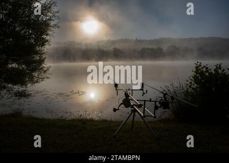 Angelruten am Ufer eines Nebelsees bei Sonnenaufgang Stockfoto