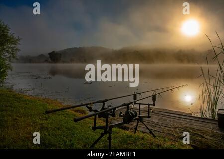 Angelruten am Ufer eines Nebelsees bei Sonnenaufgang Stockfoto