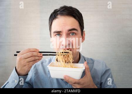 Ein junger Mann in einem blauen Hemd isst Nudeln aus einer Schachtel mit einem unzufriedenen Gesicht. Mittagessen im Büro. Geschmackloses Junkfood Stockfoto