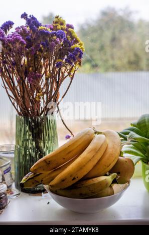 Teller mit Bananen neben Blumen auf der Fensterbank neben dem Fenster Stockfoto