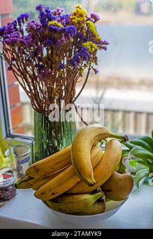 Teller mit Bananen neben Blumen auf der Fensterbank neben dem Fenster Stockfoto