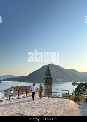 Perast, Montenegro – 03. august 2023: Mann und Frau gehen Hand vor dem Hintergrund des Glockenturms der Nikolaikirche. Perast Stockfoto