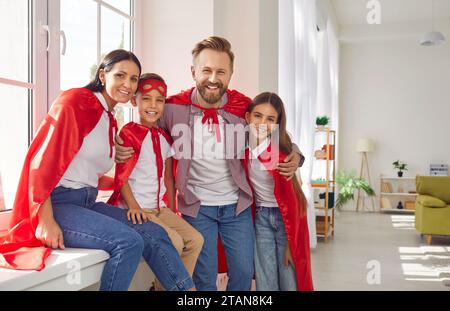 Glückliche Familie in Superheldenkostümen, die zu Hause auf der Fensterbank sitzt und in die Kamera blickt. Stockfoto