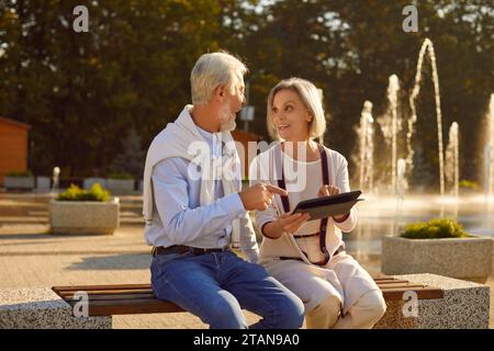 Seniorenpaar, das auf einer Bank im Park sitzt und ein digitales Tablet benutzt, um sich im Ruhestand zu fühlen Stockfoto
