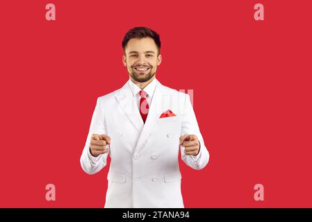 Schöner erfolgreicher glücklicher Mann, der lächelt und mit den Fingern auf dich zeigt, isoliert auf rotem Atelierhintergrund. Stockfoto