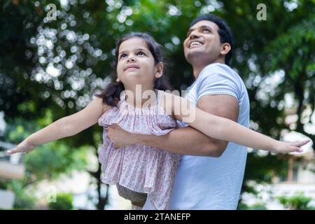 Glücklicher indischer Vater, der die Tochter im Arm hält und fliegendes Flugzeug spielt, und die gemeinsame Freizeit im Garten oder Park genießt. Kinderträumkonzept Stockfoto