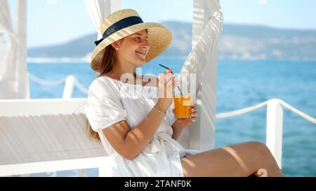 Eine Frau mit Strohhut setzt sich in einem Pavillon auf einem hölzernen Seebrück nieder. Sie schlürft an ihrem Drink und sieht die Ruhe und das Meer, verkörpert Luxusreisen und Sommer Stockfoto