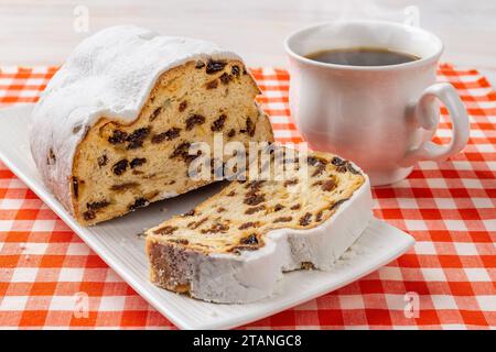 Süße Stollenscheibe und Tasse heißen Tee. Ein Stück traditionelles deutsches Weihnachtsbrot auf einem Teller und eine Tasse Kaffee auf einem rot karierten Hintergrund. Stockfoto