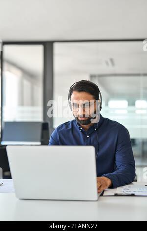 Indian man Customer Support Manager trägt ein Headset und arbeitet im Büro an einem Laptop. Stockfoto