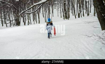 Happy Boy trägt seine Plastikschlitten den Hügel hinauf, begeistert vom Schnee und all den Outdoor-Aktivitäten, die er während der Wintersaison bringt Stockfoto